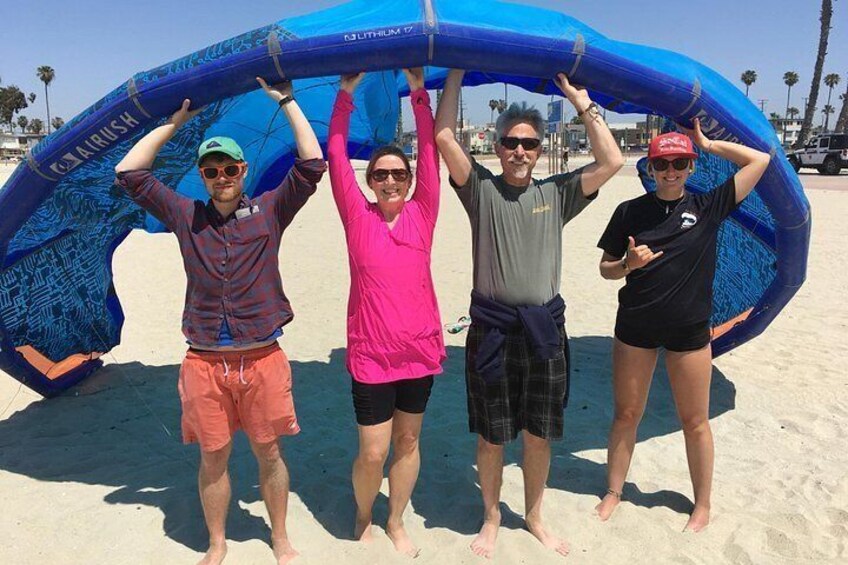 Four people holding a blue inflatable structure on a sandy beach with palm trees in the background. 3