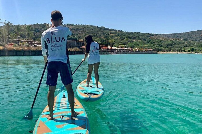 Two people paddleboarding on turquoise water with a hilly landscape in the background. 1