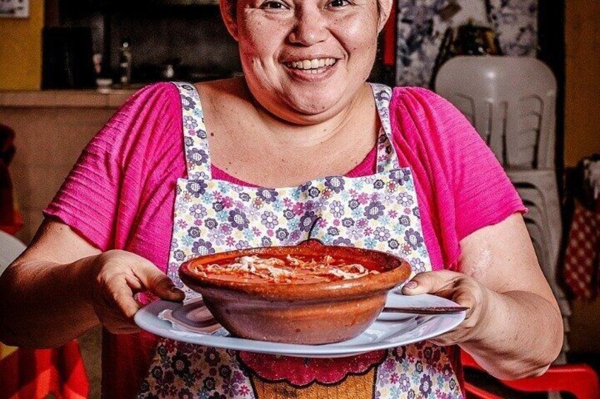 A person holding a large clay pot with a pizza inside, wearing a floral apron. 4