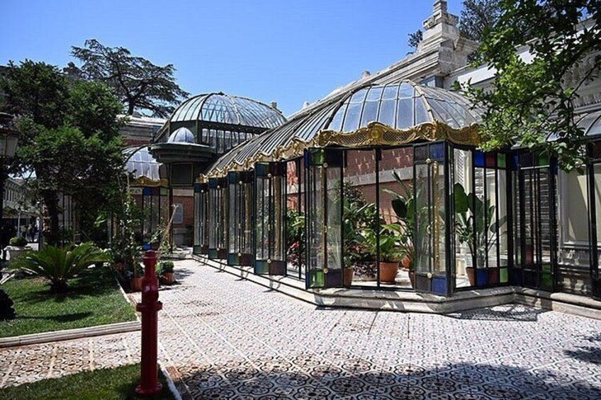 A greenhouse with glass walls and a tiled floor, surrounded by potted plants and trees. 1