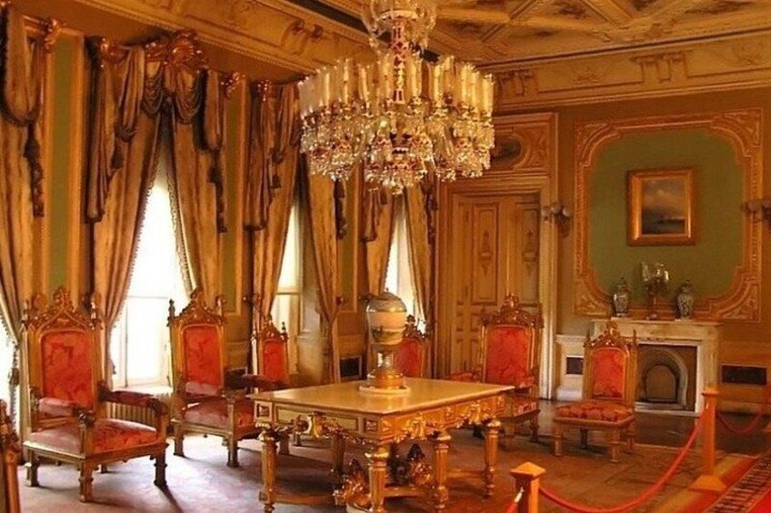 An ornate room with a large chandelier, red upholstered chairs, and a fireplace. 4