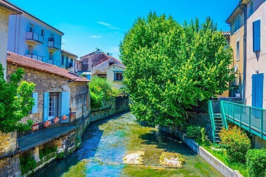 A narrow waterway flanked by stone buildings with blue shutters, a large tree, and a clear blue sky. 4