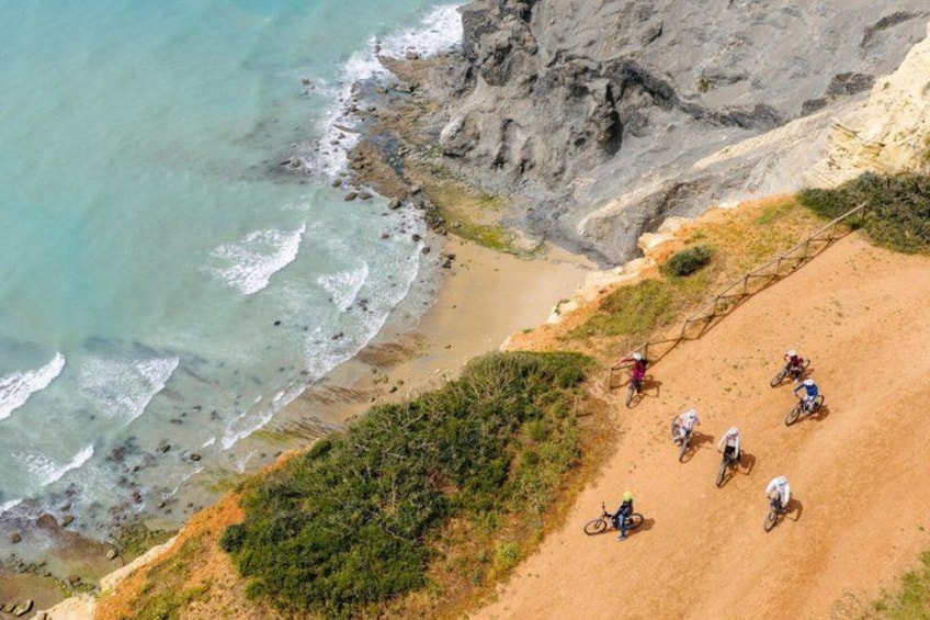 Un grupo de ciclistas en un sendero de tierra cerca de una playa con acantilados rocosos y olas del océano. 3