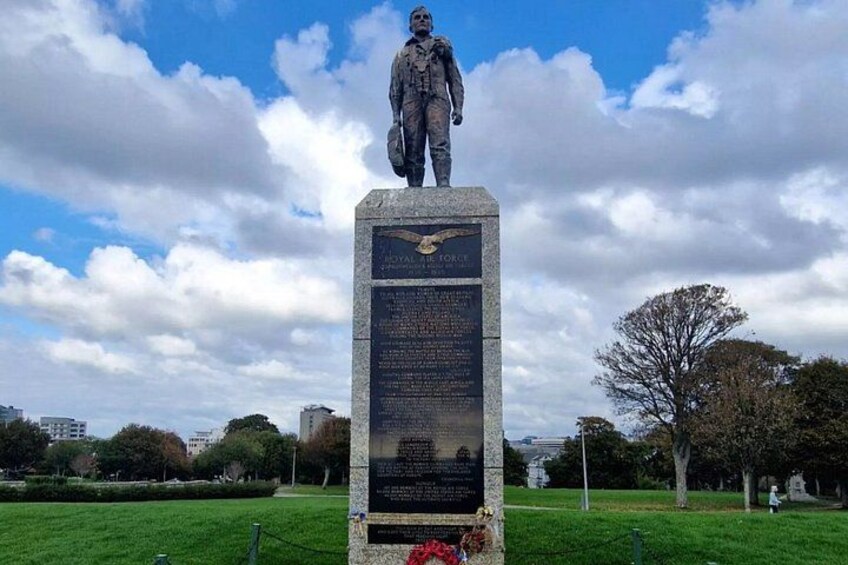 A statue on a pedestal with a plaque, set in a park with trees and a cityscape in the background. 4
