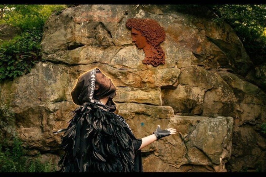 A person in a black feathered hat and black outfit standing in front of a rock formation with a carved face. 3
