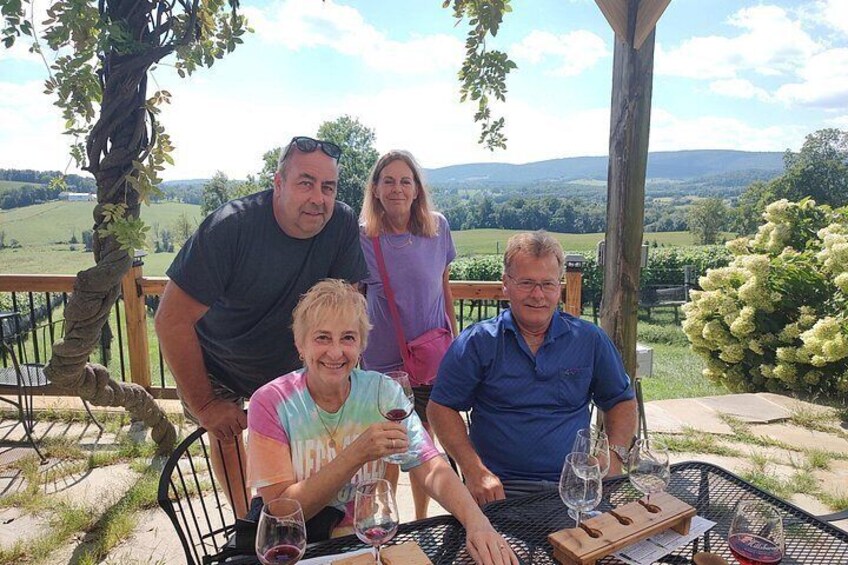 A group of people seated at an outdoor table with wine glasses, overlooking a scenic landscape with trees and hills. 5