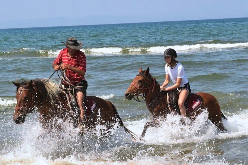 Two people riding horses on a beach with waves crashing around them. 1