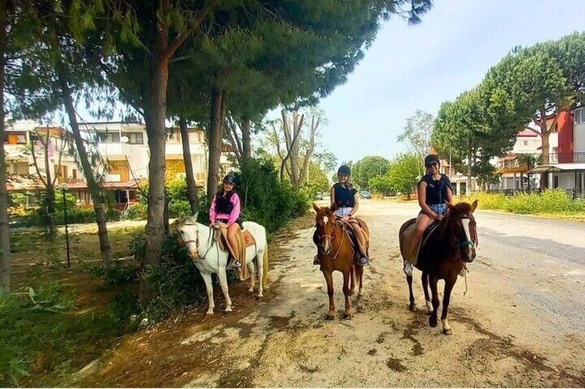 Three people riding horses on a dirt road with trees and buildings in the background. 2