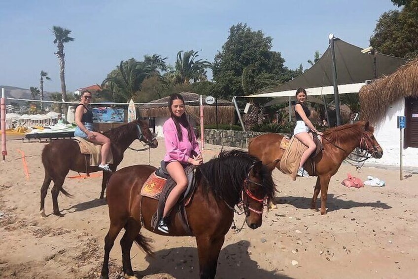 Three people riding horses on a sandy beach with palm trees and a thatched-roof hut in the background. 5
