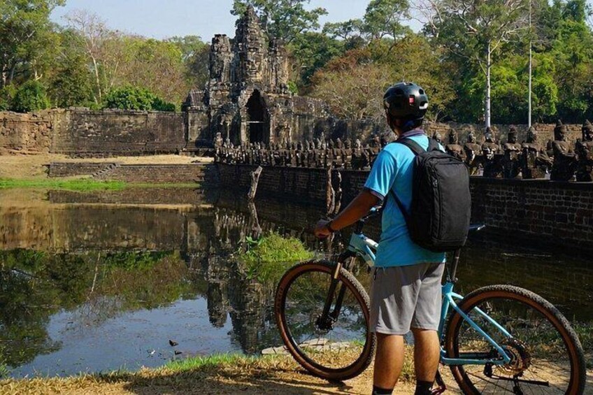 Una persona está junto a un cuerpo de agua, con una bicicleta, y un edificio de piedra al fondo, rodeado de vegetación. 1