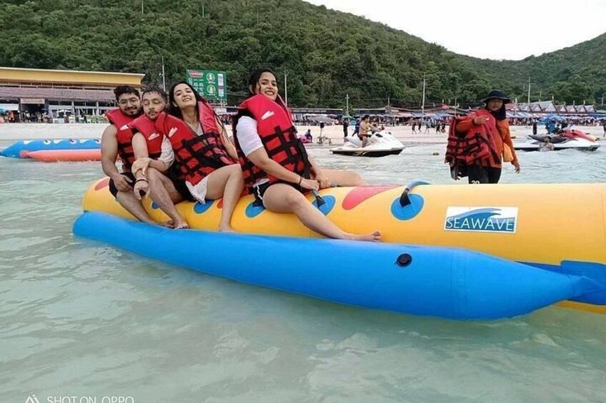 People on a yellow inflatable raft with a blue stripe, wearing red life jackets, on a body of water with boats in the background. 5