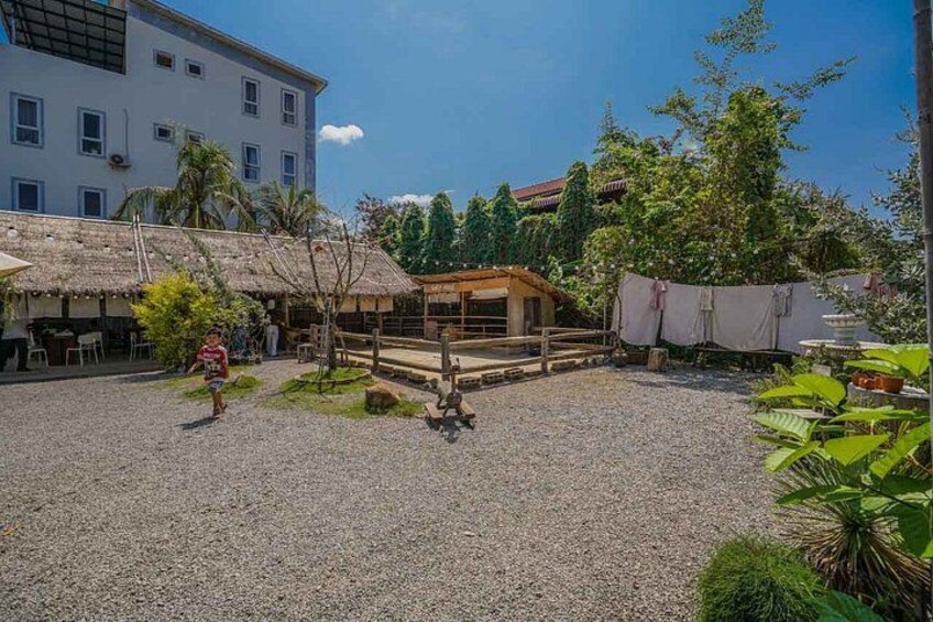 Un patio con una estructura de techo de paja, un edificio blanco y vegetación tropical. 2