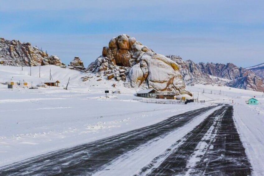 A snow-covered landscape with a large rock formation, a paved road, and a small building. 2