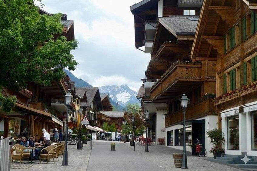 A cobblestone street lined with wooden buildings, outdoor seating, and a mountain backdrop. 5