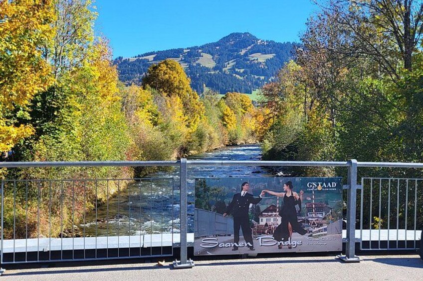 A scenic view of a river with autumn foliage, a bridge, and a sign for 'Saaren Bridge' with a 'GSTAAD' logo. 1