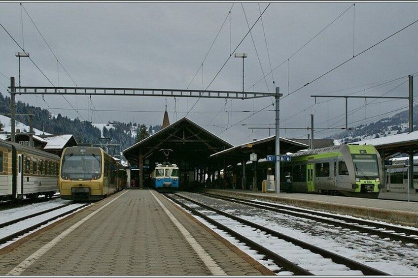 A train station with multiple tracks and trains, snow on the ground, and a mountainous backdrop. 2