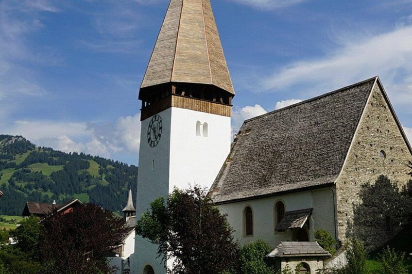 A church with a clock tower, surrounded by trees and houses, set against a mountainous backdrop. 4