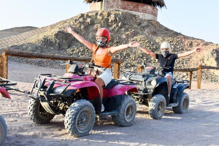 Two people on ATVs on a sandy terrain with a rocky hill in the background. 2