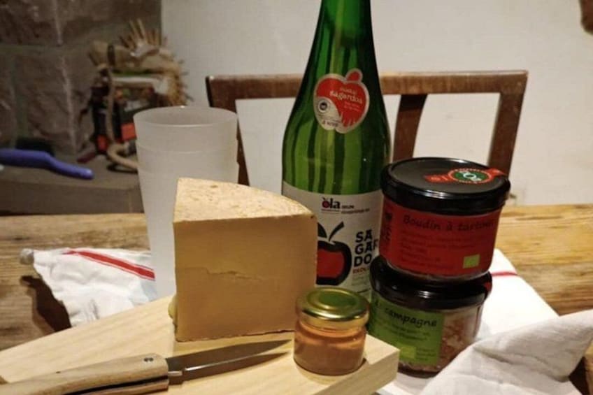 A wooden cutting board with a knife, a block of cheese, a bottle of olive oil, a jar of mustard, and a container of bread. 3