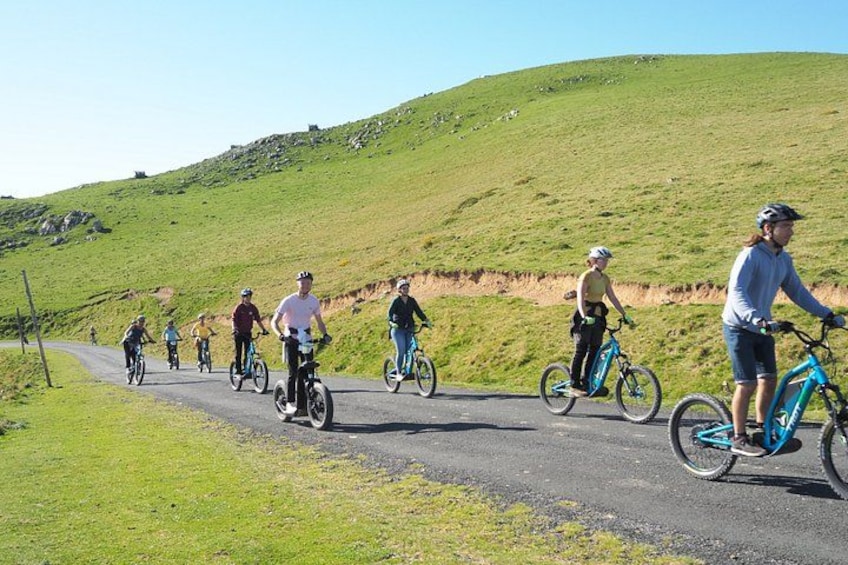 A group of cyclists on a paved road with a grassy hill in the background. 2