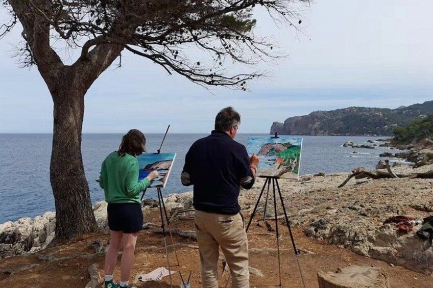 Two people painting by the seaside with a tree and rocky terrain in the background. 4