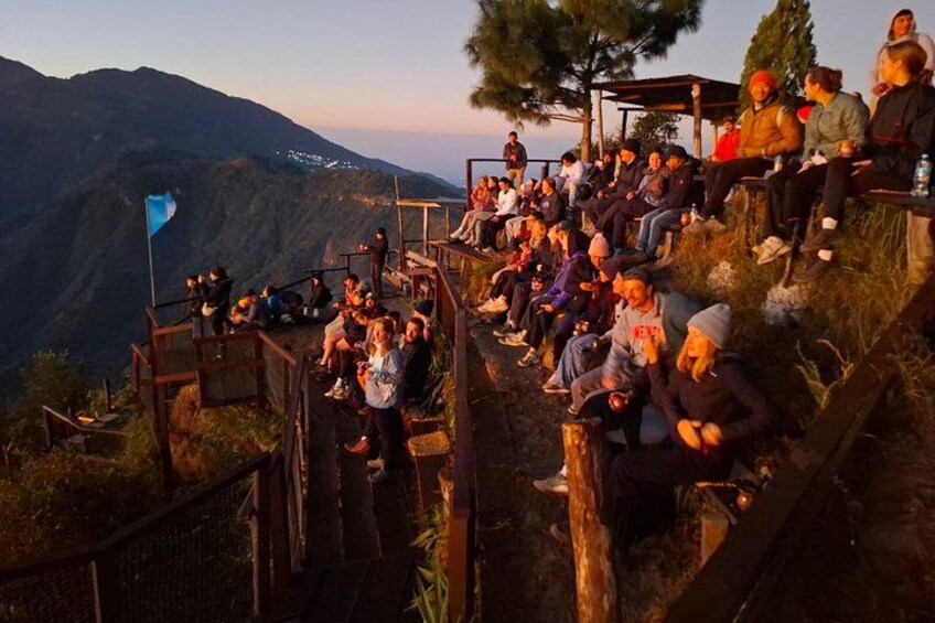 A group of people sitting on wooden benches on a hillside, overlooking a mountainous landscape. 5