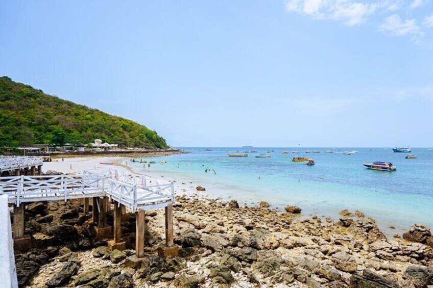 A rocky beach with a wooden pier, clear turquoise water, and boats in the distance. 5