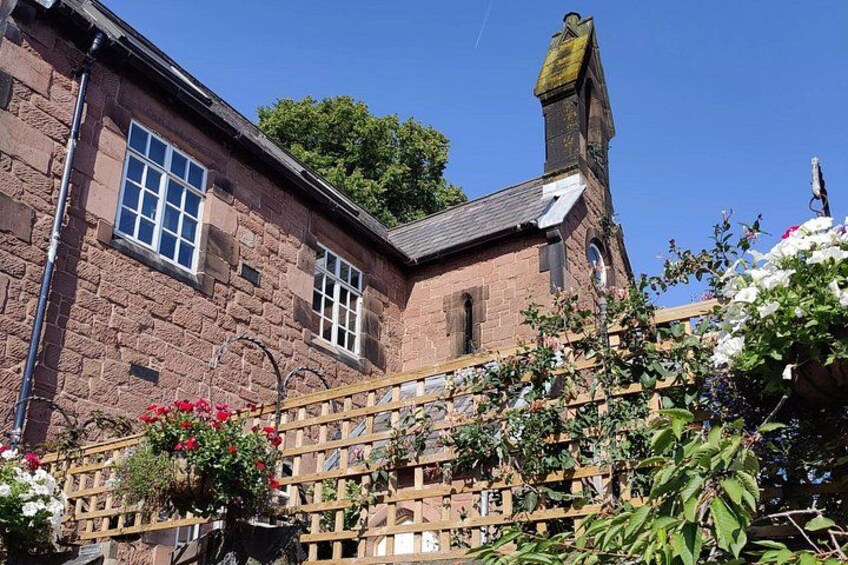 A stone building with a clock tower, surrounded by flowering plants and a wooden fence. 1