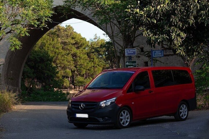 A red car parked under a stone bridge with trees and a building in the background. 4