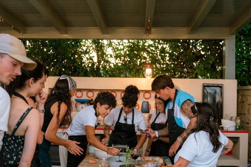 A group of people engaged in a cooking activity outdoors, with a rustic kitchen setup and greenery in the background. 5