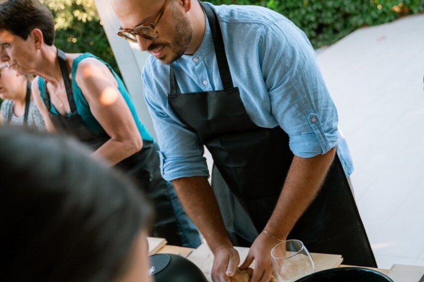 A person preparing food outdoors, wearing a denim shirt and apron, with greenery in the background. 2