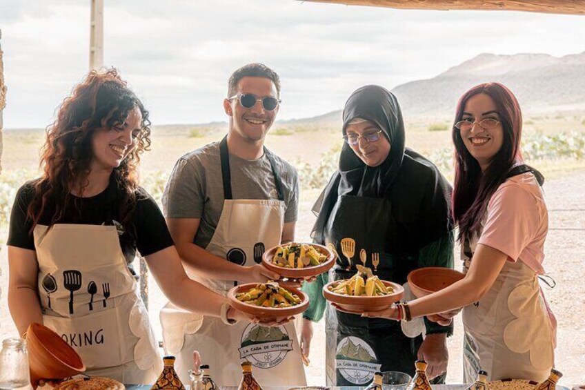 Four people wearing aprons with cooking utensils, holding plates of food, and standing outdoors with a desert landscape in the background. 5