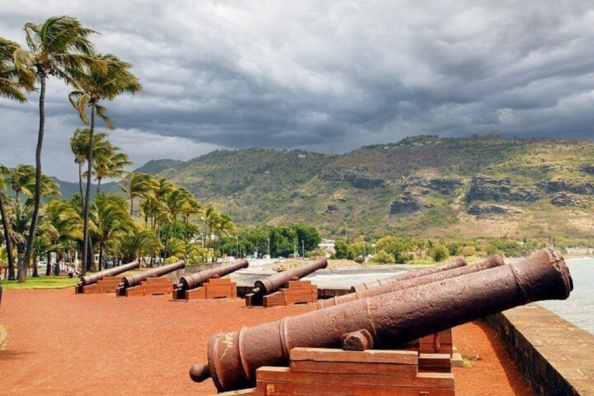 A coastal area with a row of cannons lined up along a walkway, palm trees, and a mountainous backdrop under a cloudy sky. 3