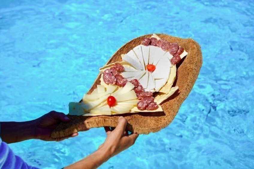 A person holding a wooden board with sliced cheese, salami, and a cherry tomato by a pool. 2