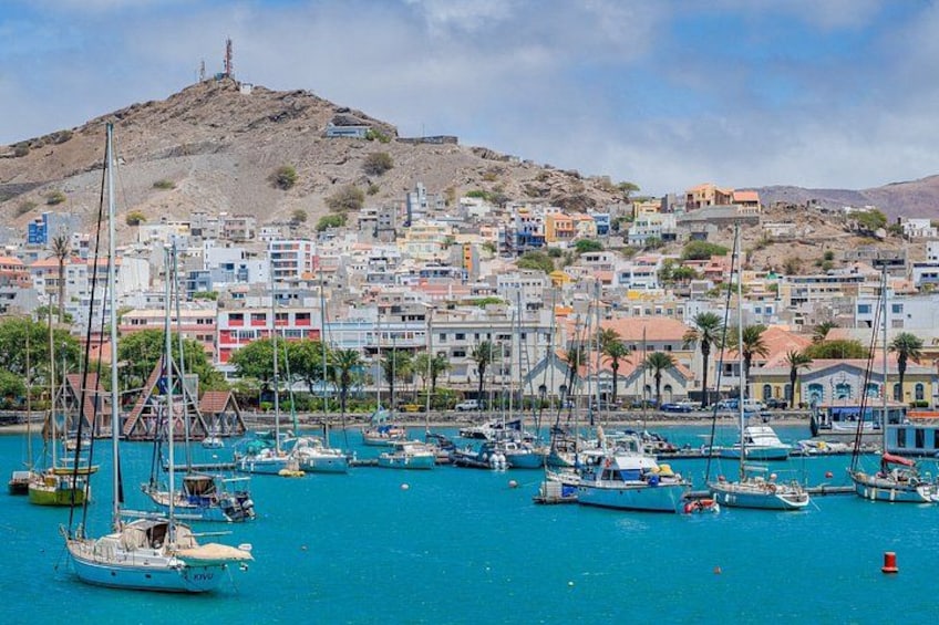 A coastal town with a marina filled with boats, a prominent hill in the background, and colorful buildings. 2