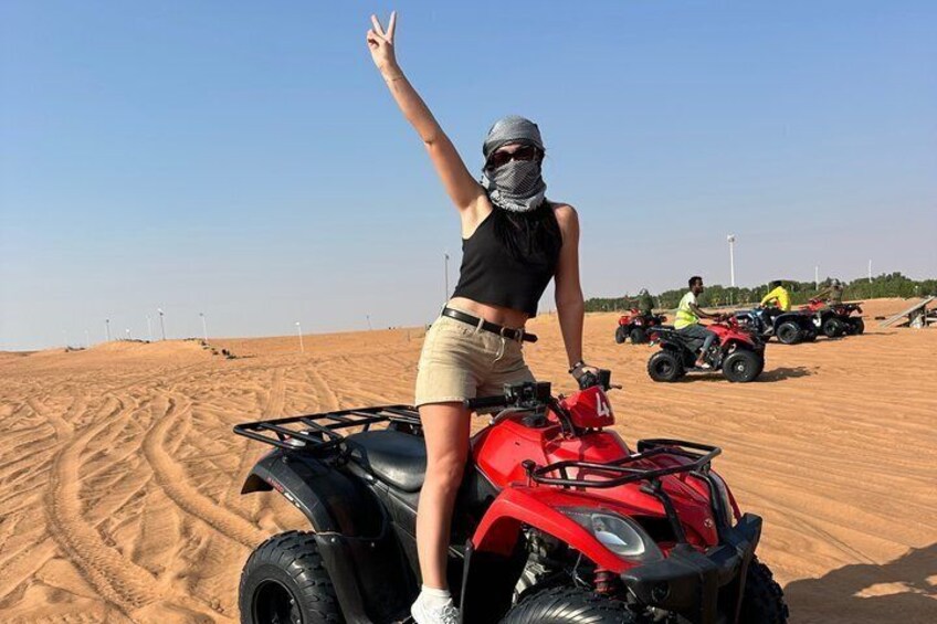 A person on a red quad bike in a desert with sand dunes and other quad bikes in the background. 1