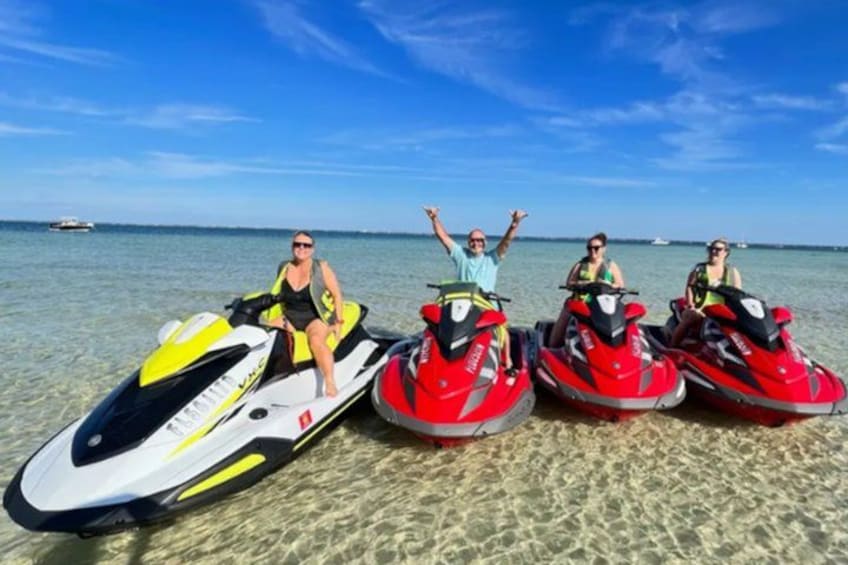 Four jet skis on a sandy beach with clear water and a boat in the background. 2