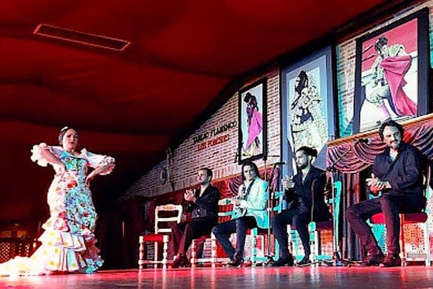 A stage with a performer in a colorful dress, seated audience, and framed artwork on the wall. 2