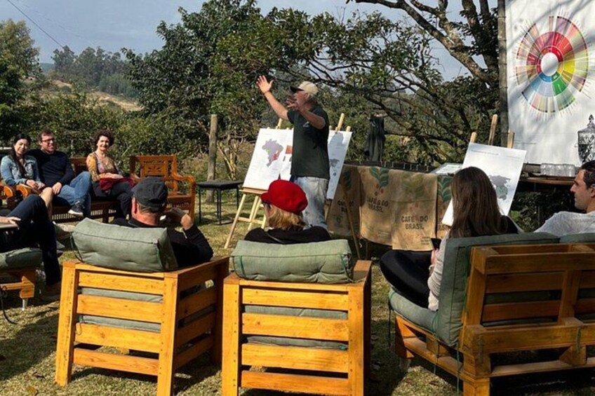A group of people sitting on wooden chairs outdoors, some facing an artist painting on a canvas. 5