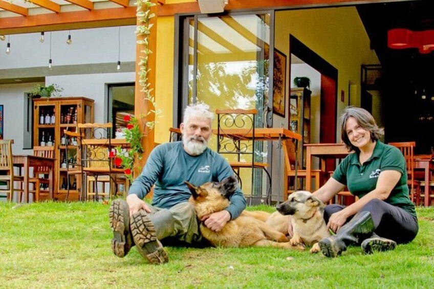 A man and a woman sitting on grass with two dogs in a patio area with wooden furniture and a glass door. 5