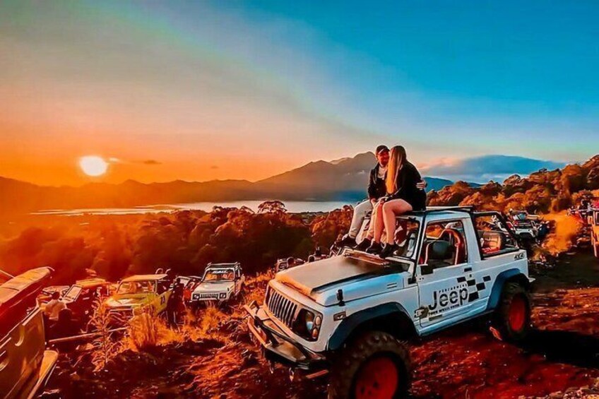 A couple on a white Jeep with 'Jeep' written on the side, parked on a rocky terrain with a sunset in the background. 5