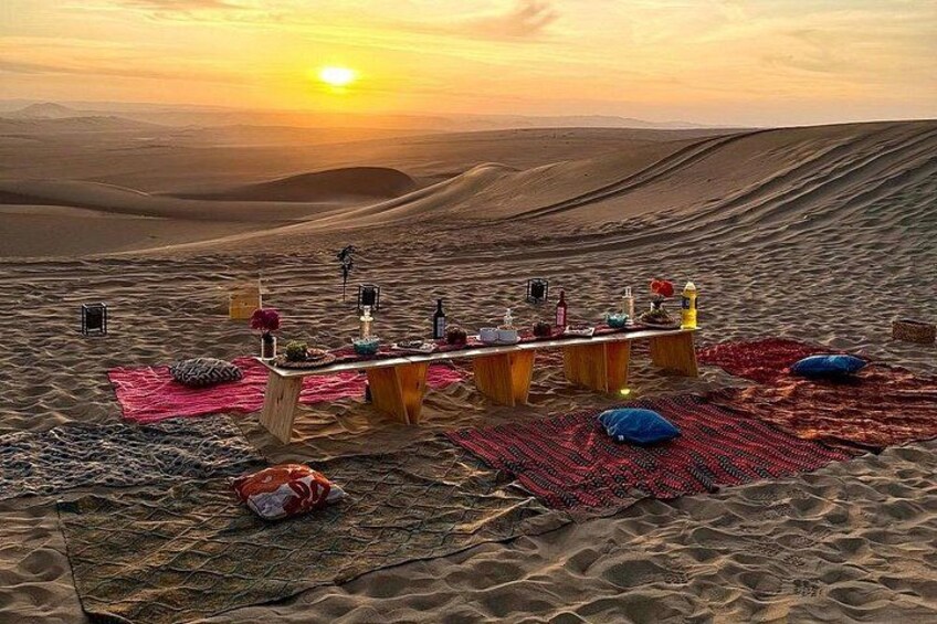 Un pique-nique organisé dans le désert, avec une table, des couvertures, des coussins et un coucher de soleil en arrière-plan. 1
