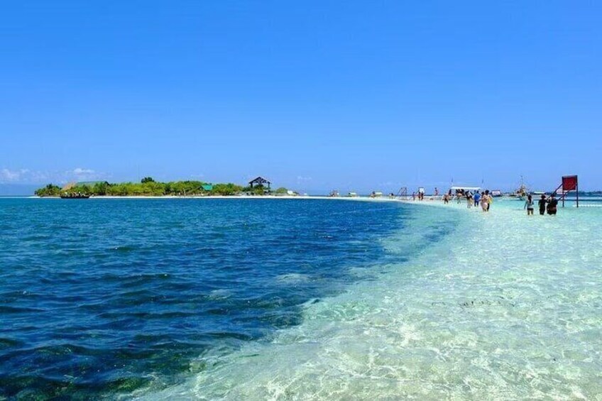 A beach with clear turquoise water, a small island with buildings, and people walking along the shore. 3