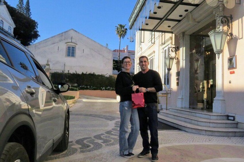Two people standing in front of a building with a car parked nearby. 2