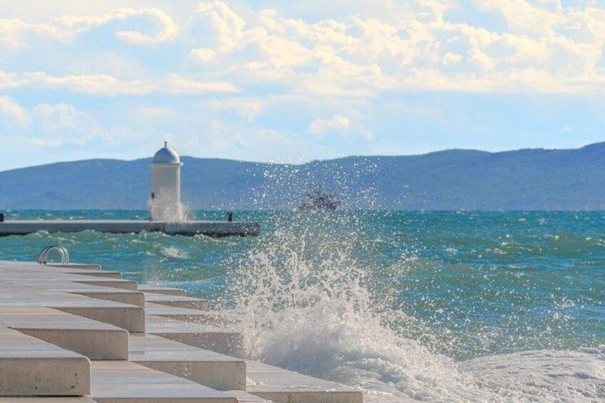 A coastal scene with a lighthouse, waves crashing onto a pier, and mountains in the background. 5