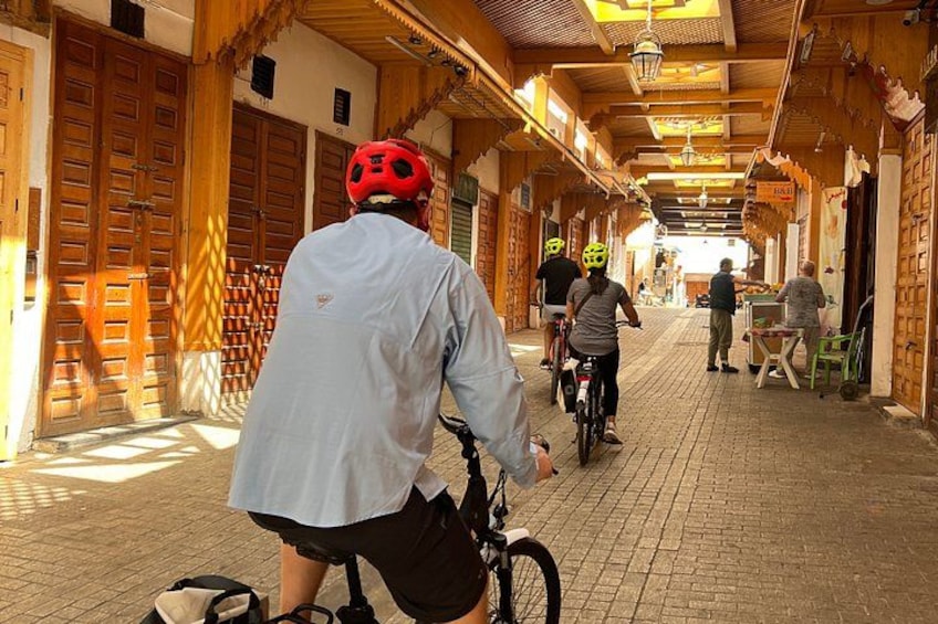 A person on a bicycle wearing a red helmet, riding down a cobblestone street with wooden doors and a covered walkway. 4