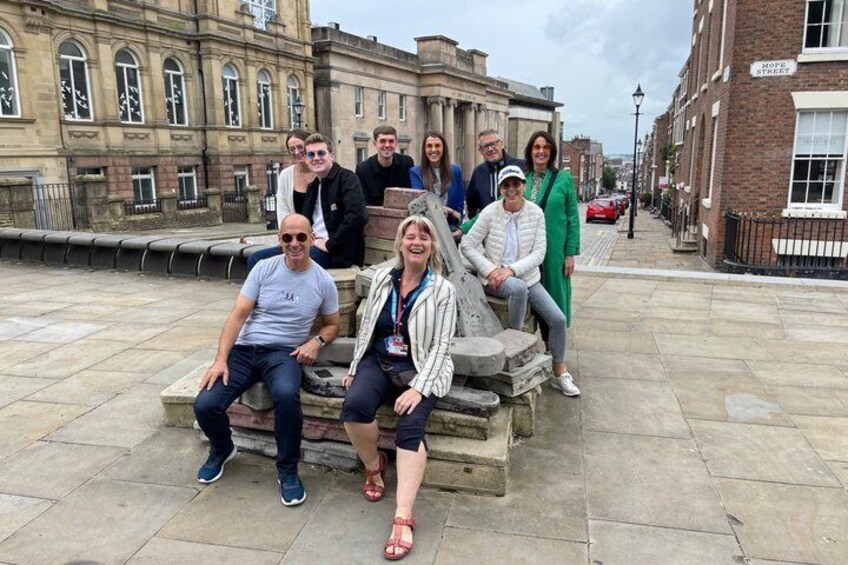 A group of people sitting on a stone bench in an urban setting with historic buildings in the background. 2