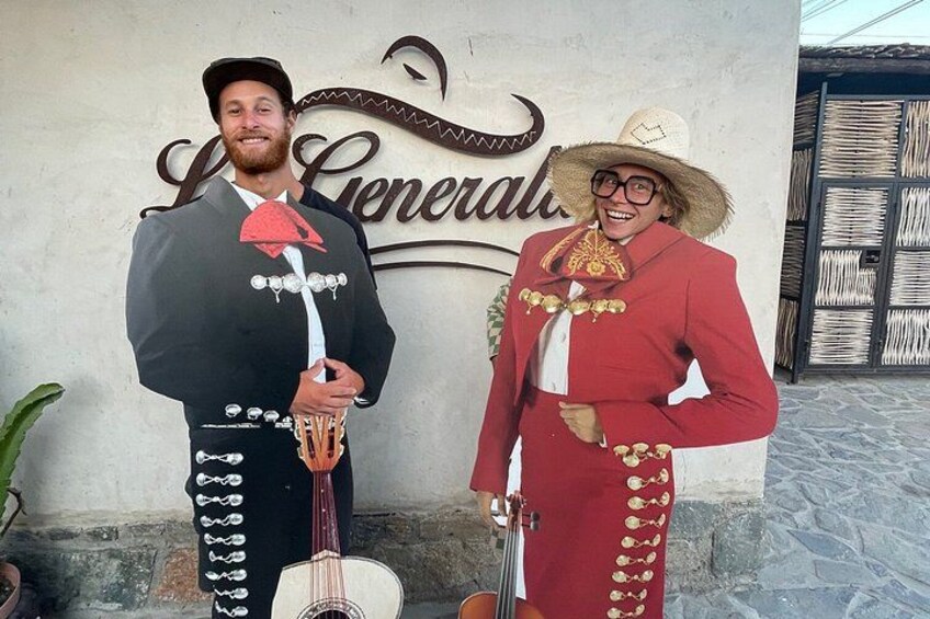 Two people in traditional Mexican attire, one holding a guitar and the other a violin, standing in front of a building with a sign. 5