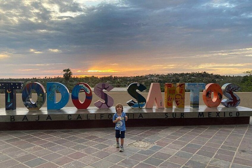 A person standing in front of a sign with colorful letters spelling 'TODOS SANTOS' and 'BAJA CALIFORNIA SUR MEXICO' on a tiled floor. 3