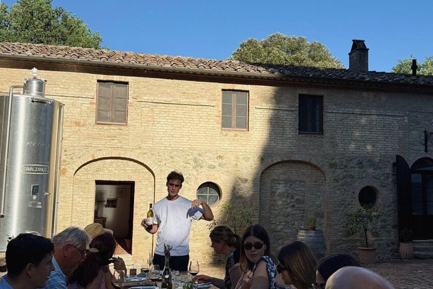 A man standing in front of a rustic building with a group of people seated at a table. 1